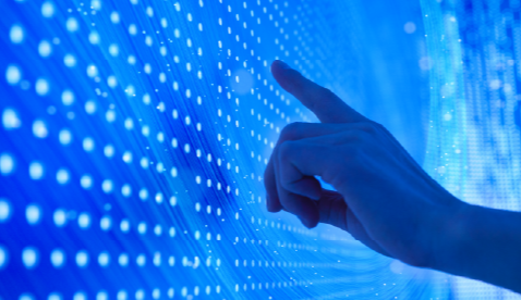 Mano interactuando con una pantalla digital azul compuesta por filas de puntos blancos, simulando una interfaz tecnológica o de datos. Representa la conexión entre el usuario y sistemas digitales en entornos de innovación.